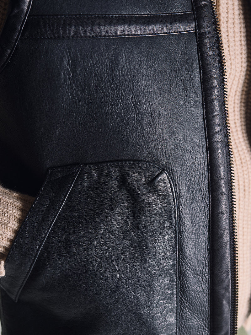 Close-up of the Blue de Gênes Saga Shearling Gilet - Scarab, highlighting the texture of German Merino sheepskin, a visible zipper, and a beige knitted sweater sleeve.