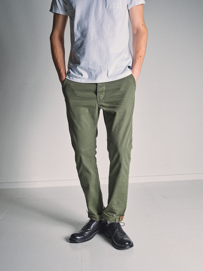 A person stands indoors against a light background, wearing a white short-sleeve shirt, Blue de Gênes Paulo Pavia 1 V2 Chino - Army pants in cotton stretch fabric, and black shoes, with hands in pockets and head out of frame.