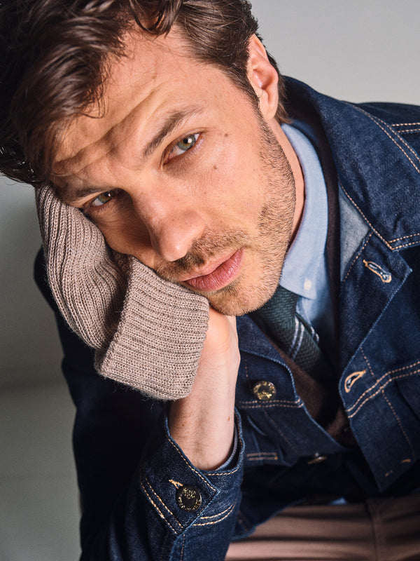 A man with tousled brown hair leans his face on his hand, looking up. He wears a Blue de Gênes denim jacket over a collared shirt and the Blue de Gênes Koi Wool Gilet in Chocolate, its sleeve covering his hand.