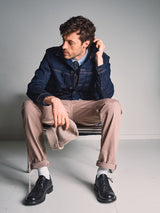 A man with curly hair sits on a chair, wearing a denim jacket, light blue shirt, Blue de Gênes Paulo Pavia 1 V2 Chino - Fungi pants, white socks, black shoes, and holding a hat while looking to the side.