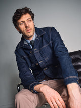 A man with tousled hair sits on a black leather chair, sporting the Blue de Gênes Nekutaki Tie #D - Green Stripe over a shirt and denim jacket, paired with beige pants. He gazes thoughtfully to the side in a minimalist setting.