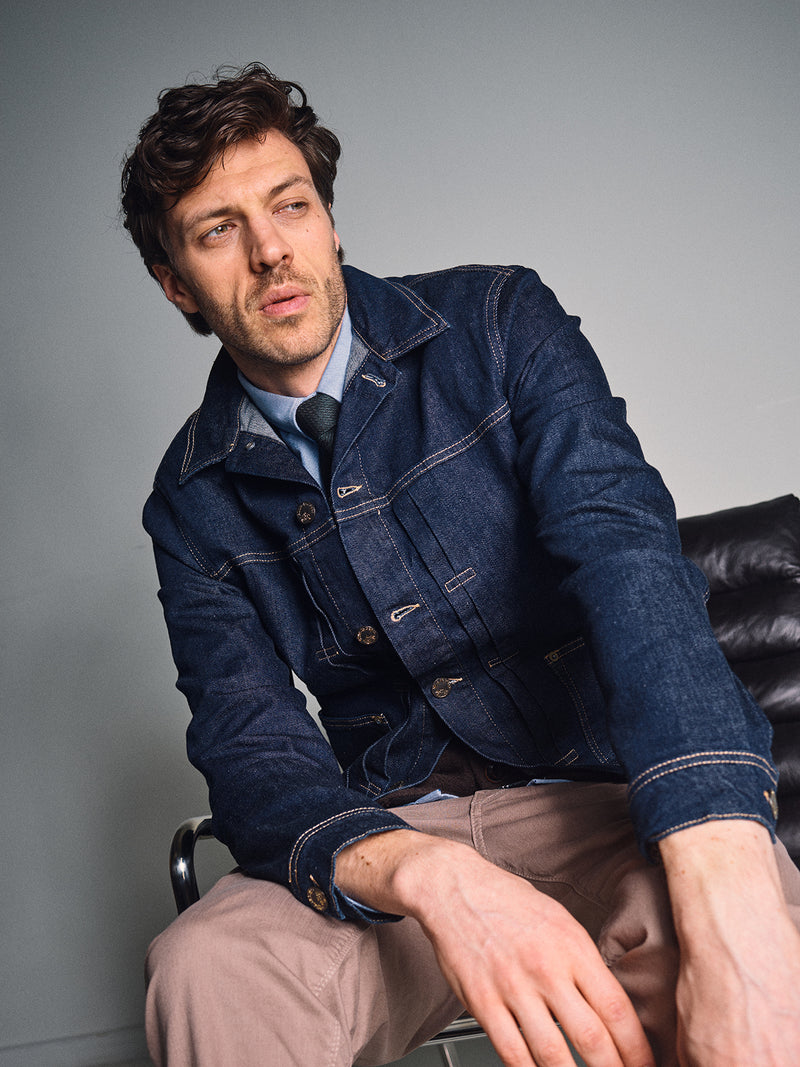 A man with tousled hair sits on a black leather chair, sporting the Blue de Gênes Nekutaki Tie #D - Green Stripe over a shirt and denim jacket, paired with beige pants. He gazes thoughtfully to the side in a minimalist setting.