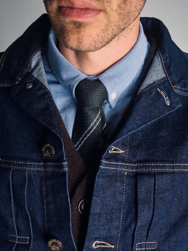 A close-up of a man in a blue denim jacket, blue shirt, brown vest, and the Nekutaki Tie #D - Green Stripe by Blue de Gênes. Cropped below his nose, the image highlights this stylish mens accessory and outfit.