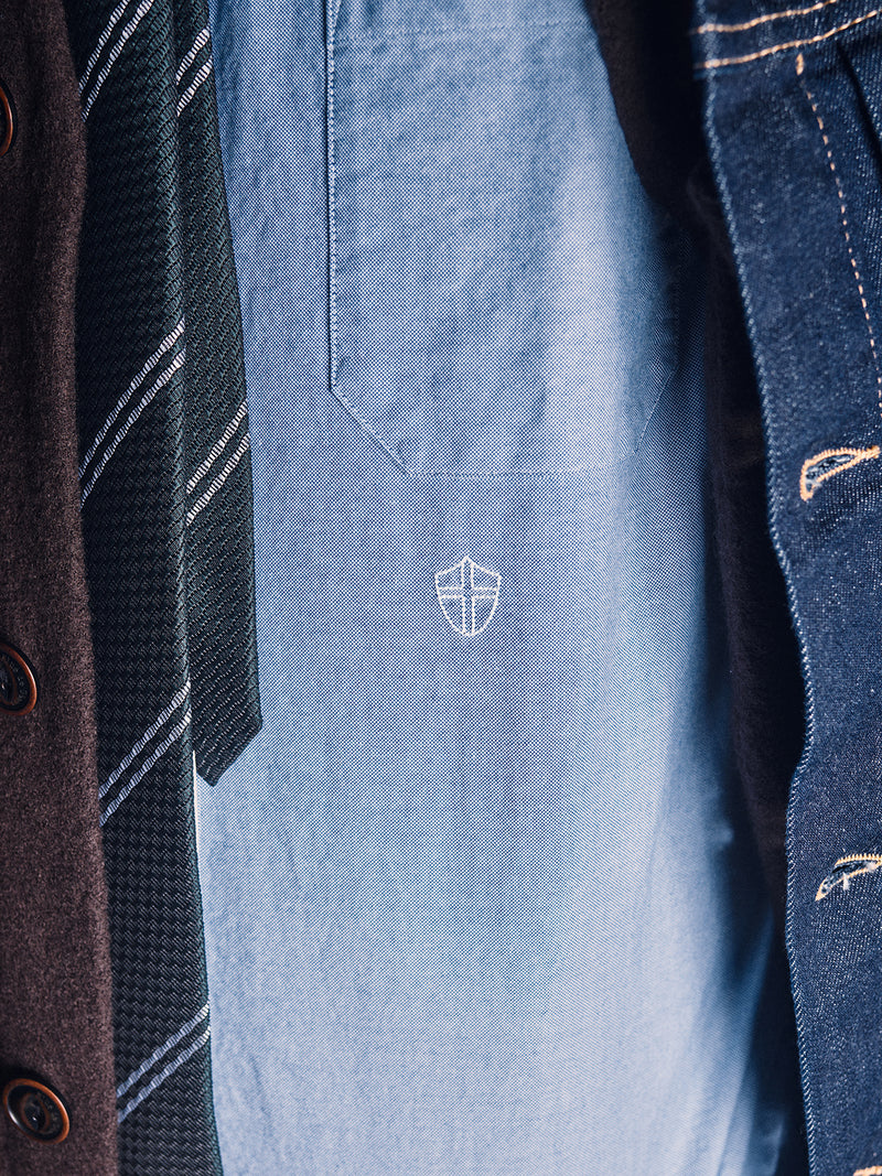 Close-up of a person wearing a blue shirt with a white embroidered shield, Nekutaki Tie #D - Green Stripe by Blue de Gênes, brown coat, and denim jacket. Only the chest area is shown.