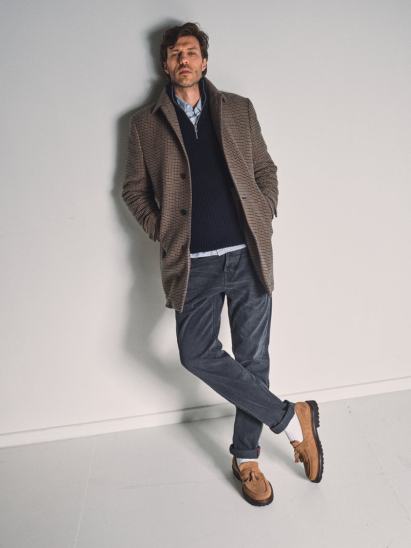 A man leans against a white wall with his hands in his pockets, wearing a brown plaid coat, navy sweater, light blue shirt, Blue de Gênes Giacomo BLK Used Jeans in grey denim, and tan loafers.