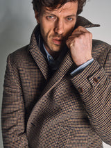 A man with tousled hair wears the Blue de Gênes Massone Check Coat - Multi Check, holding up the collar with one hand and looking seriously at the camera against a plain background.