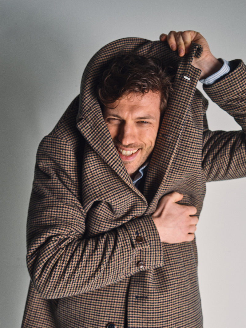 A man smiles playfully while pulling the collar of his Blue de Gênes Massone Check Coat - Multi Check over his head, partially covering his face. He stands against a plain, light background.