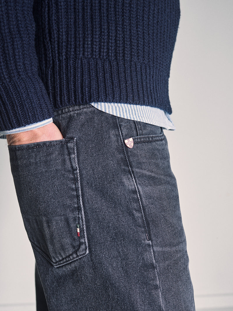 A person wearing a navy blue knit sweater, a blue-and-white striped shirt, and Blue de Gênes Giacomo BLK Used Jeans in grey denim with one hand in their pocket, photographed from the side.