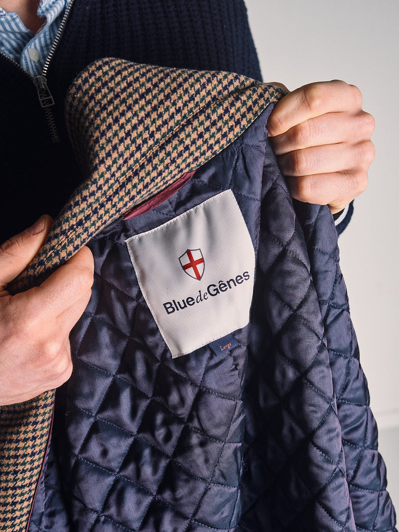 A person holds open the Massone Check Coat - Multi Check by Blue de Gênes, revealing the inside label with the Blue de Gênes name and a red and white shield logo. The coat features a houndstooth pattern on the collar.