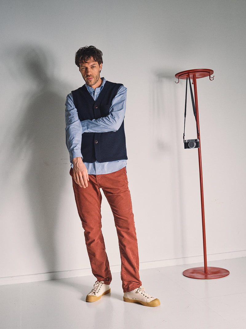 A man in a Blue de Gênes Koi Wool Gilet - Navy, striped shirt, and red pants stands indoors on a white floor beside a red coat rack with a camera. He appears relaxed, with one arm bent and his head slightly tilted.