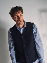A man with curly brown hair models the Blue de Gênes Koi Wool Gilet in Navy, standing against a light background and gazing thoughtfully to the side.