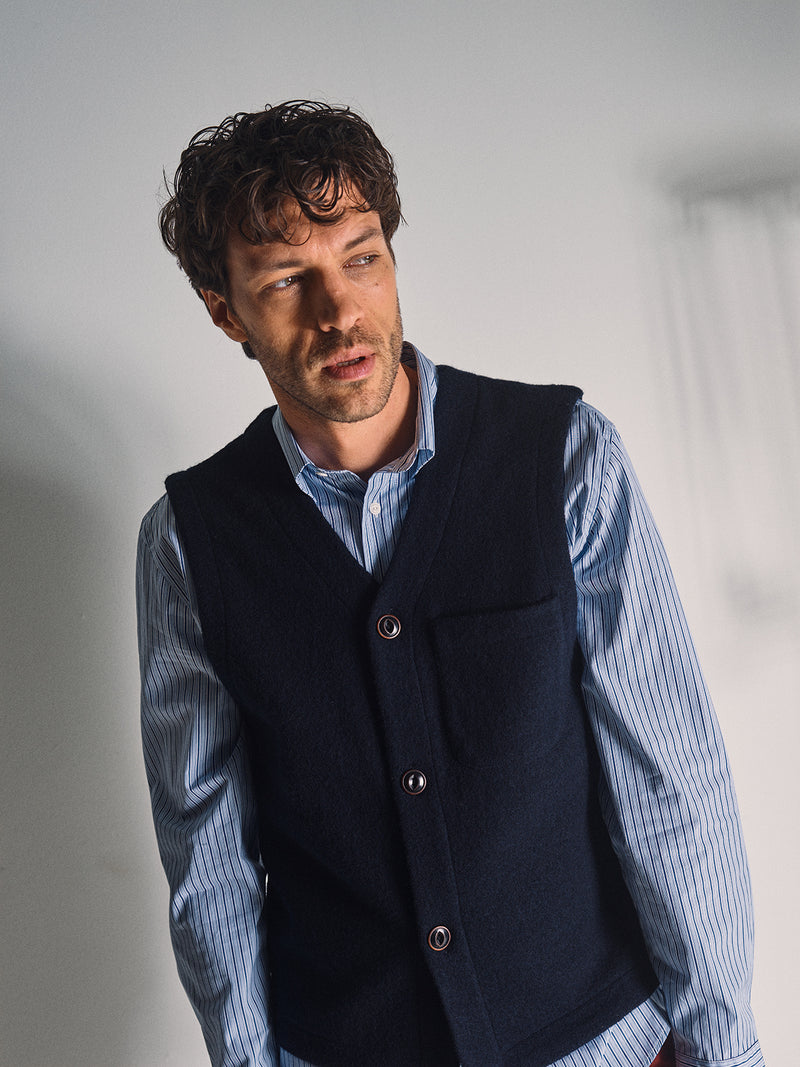 A man with curly brown hair models the Blue de Gênes Koi Wool Gilet in Navy, standing against a light background and gazing thoughtfully to the side.