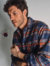 A man with wavy brown hair and a beard wears the Blue de Gênes Yodo Check Overshirt - Multi Check. He looks to the side with a serious expression, hands together, with part of a sign visible behind him.