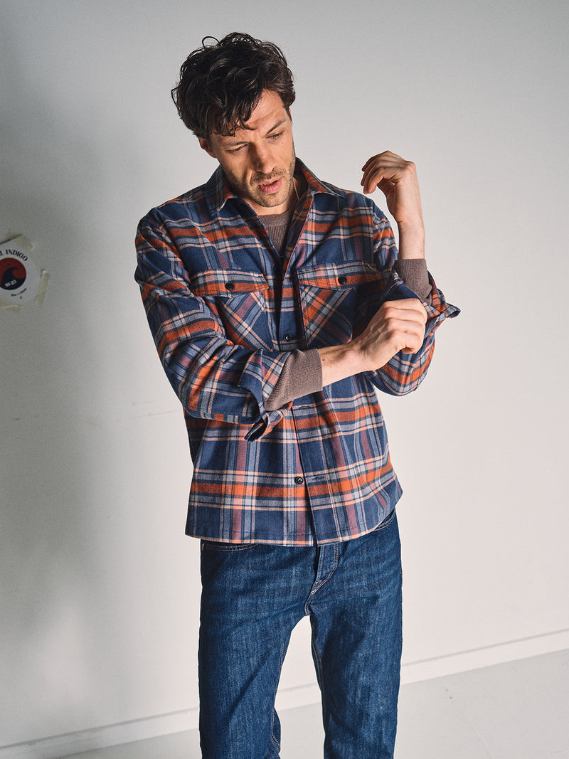 A man stands indoors in relaxed fit jeans and a Blue de Gênes Yodo Check Overshirt - Multi Check, looking down as he rolls up his left sleeve. The soft-touch moleskin fabric offers comfort against a plain background with a sticker on the wall.
