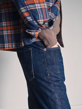 A person wearing a blue and orange plaid shirt over a brown sweater has their hand in the pocket of Blue de Gênes Giacomo Dark Horse Jeans - Denim Blue, standing against a plain background.