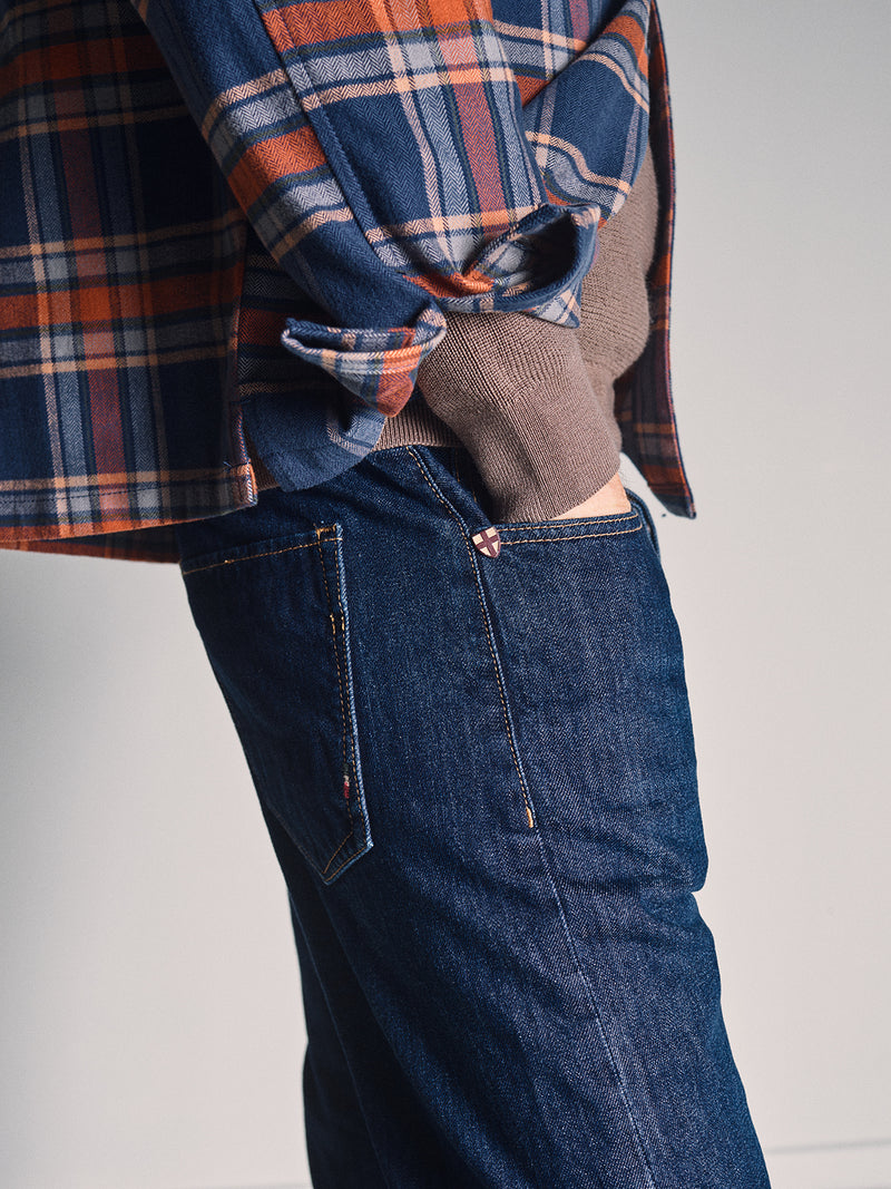 A person wearing a blue and orange plaid shirt over a brown sweater has their hand in the pocket of Blue de Gênes Giacomo Dark Horse Jeans - Denim Blue, standing against a plain background.