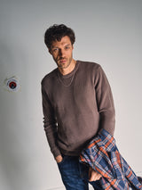 A man with wavy dark hair wears the Blue de Gênes Porto Knit - Fungi, a brown 100% wool crewneck sweater, paired with blue jeans and a silver chain. He stands against a plain wall, holding a plaid jacket and looking at the camera neutrally.