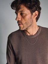 A man with wavy brown hair and light stubble wears the Blue de Gênes Porto Knit - Fungi sweater and a thin chain necklace, gazing thoughtfully to his left against a plain light background.