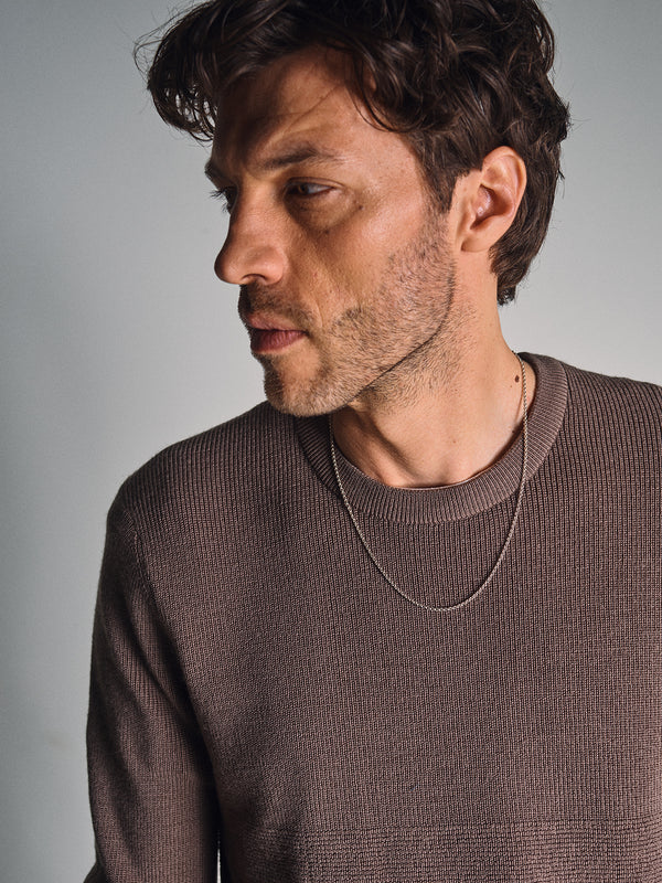 A man with wavy brown hair and light stubble wears the Blue de Gênes Porto Knit - Fungi sweater and a thin chain necklace, gazing thoughtfully to his left against a plain light background.
