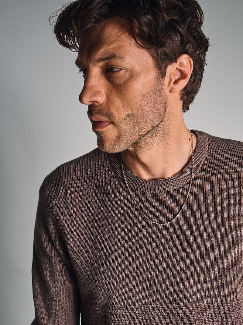 A man with wavy brown hair and light stubble wears the Blue de Gênes Porto Knit - Fungi sweater and a thin chain necklace, gazing thoughtfully to his left against a plain light background.
