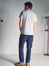 A man faces away from the camera in a white t-shirt, brown shoes, and Blue de Gênes Giacomo Dark Horse Jeans in Denim Blue. He stands in a minimalist room with a red coat rack holding a camera and a sticker on the wall.