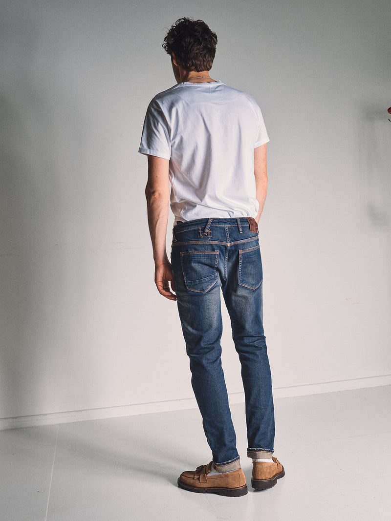 A man with short hair faces away in a white t-shirt, brown shoes, and Blue de Gênes Repi Dark Vintage Jeans in Mid Blue Denim, standing in a plain, well-lit room with a light gray wall and white floor.