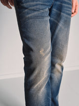 A person wears Blue de Gênes’ Repi Dark Vintage Jeans - Mid Blue Denim, a slim-fit style in dark vintage wash with comfort stretch and a small distressed patch on the front left thigh, standing against a plain light background.