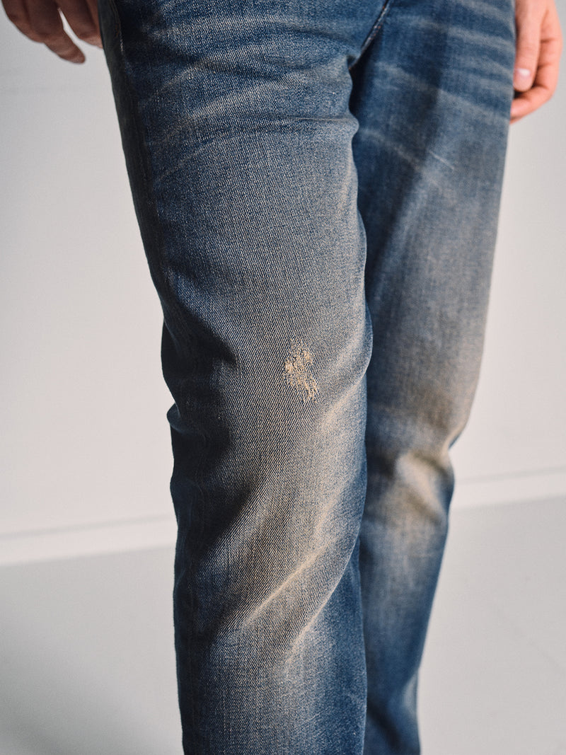 A person wears Blue de Gênes’ Repi Dark Vintage Jeans - Mid Blue Denim, a slim-fit style in dark vintage wash with comfort stretch and a small distressed patch on the front left thigh, standing against a plain light background.