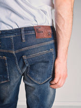 A person wears Blue de Gênes Repi Dark Vintage Jeans - Mid Blue Denim and a white t-shirt, shown waist down from the back. The jeans feature a brown leather patch with text on the waistband, and their right arm is by their side.