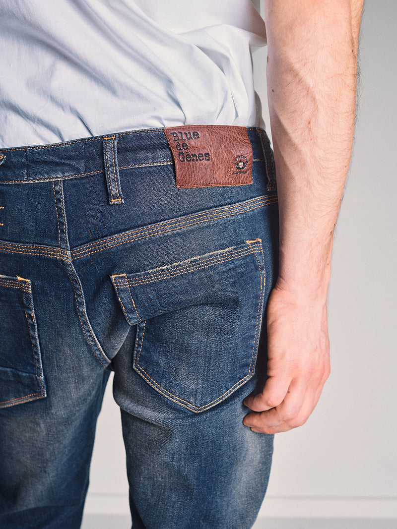 A person wears Blue de Gênes Repi Dark Vintage Jeans - Mid Blue Denim and a white t-shirt, shown waist down from the back. The jeans feature a brown leather patch with text on the waistband, and their right arm is by their side.