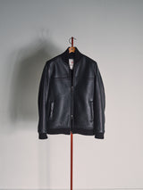 The Rufo Shearling Jacket - Scarab by Blue de Gênes, crafted from natural sheepskin with front pockets and a visible zipper, is displayed on a wooden hanger against a plain light gray wall.