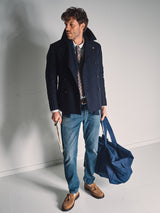 A man in a navy pea coat, patterned tie, and tan loafers stands indoors holding a folder and large navy bag. He wears Blue de Gênes Vinci Topaz Vintage Jeans - Used, crafted from comfort stretch denim for effortless style.