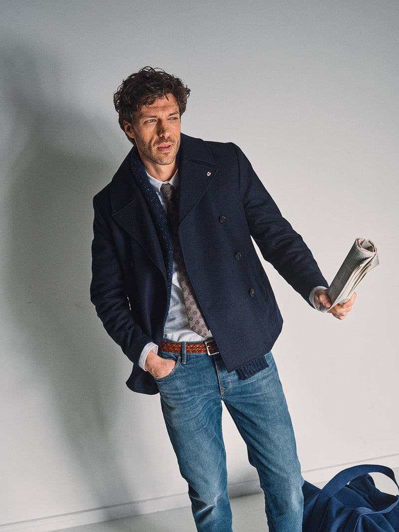 A man with wavy hair stands indoors wearing a Blue de Gênes Shiba Melton Coat in Dark Navy, a patterned tie, light shirt, blue jeans, and holds a rolled newspaper, with a blue bag on the floor beside him.