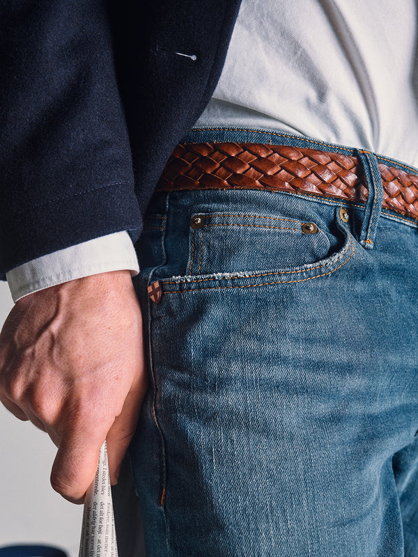 A close-up of a person wearing Blue de Gênes Vinci Topaz Vintage Jeans - Used in comfort stretch denim, paired with a woven brown leather belt, a white shirt, and a dark jacket, holding a rolled-up newspaper.