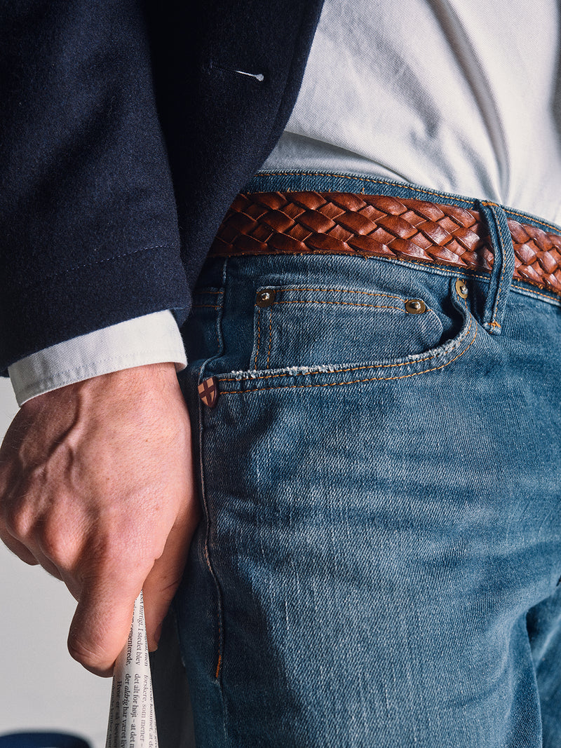 A close-up of a person wearing Blue de Gênes Vinci Topaz Vintage Jeans - Used in comfort stretch denim, paired with a woven brown leather belt, a white shirt, and a dark jacket, holding a rolled-up newspaper.