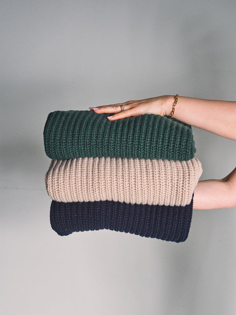 A hand holds the Blue de Gênes Yomaha Halfzip Knit sweater in Fungi, a folded, ribbed wool blend, against a plain light gray background.