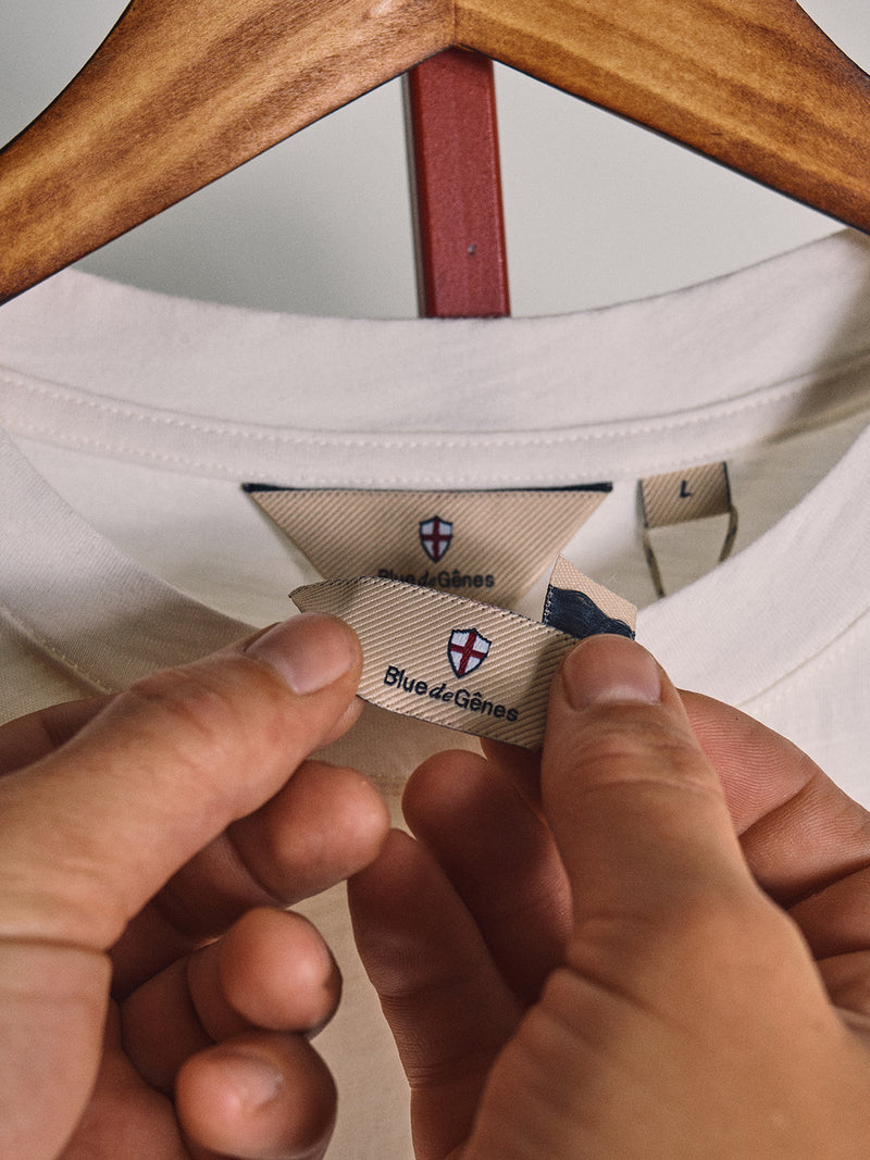 Close-up of hands holding a clothing tag on a white BGoscar T-Shirt - Snow Fall by Blue de Gênes, shown on a wooden hanger. The tag features the brand’s shield logo, regular fit, and 100% cotton fabric.