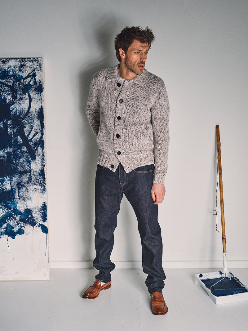 A man wearing the Blue de Gênes Sakura Knit Cardigan in Stone, dark jeans, and brown shoes stands indoors by a blue paint roller tray and a canvas splattered with blue.