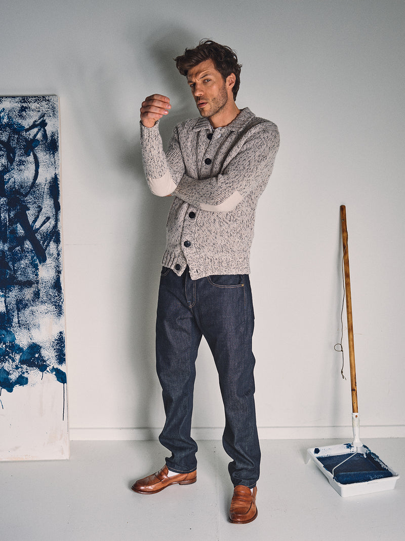 A man wearing the Blue de Gênes Sakura Knit Cardigan in Stone and jeans stands indoors, looking at his left hand. Beside him are a blue paint tray, a roller with a stick, and an abstract blue canvas leaning on the wall.
