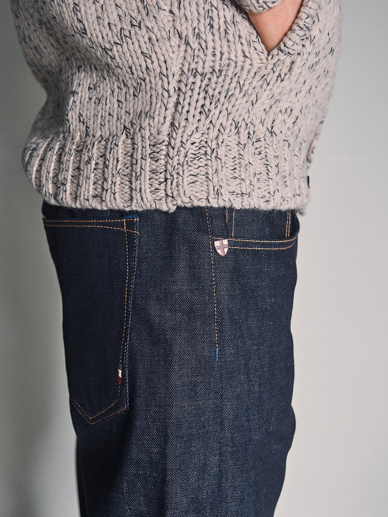 A person wearing a chunky beige knit sweater and Blue de Gênes Giacomo Selvedge V2 Jeans in raw denim stands with one hand in their pocket, photographed from the side against a plain background.