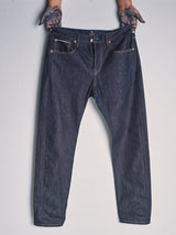 A person with tattooed hands holds up a pair of Blue de Gênes Giacomo Selvedge V2 Jeans – Raw Denim by the waistband against a light background.