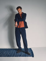 A man in a navy suit and burnt orange shirt stands on a blue rug, pointing with his right hand. He wears Blue de Gênes Etsu Cord Chino - Navy pants, highlighting their relaxed regular fit chino style.