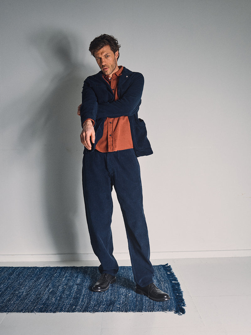 A man in a navy suit and burnt orange shirt stands on a blue rug, pointing with his right hand. He wears Blue de Gênes Etsu Cord Chino - Navy pants, highlighting their relaxed regular fit chino style.