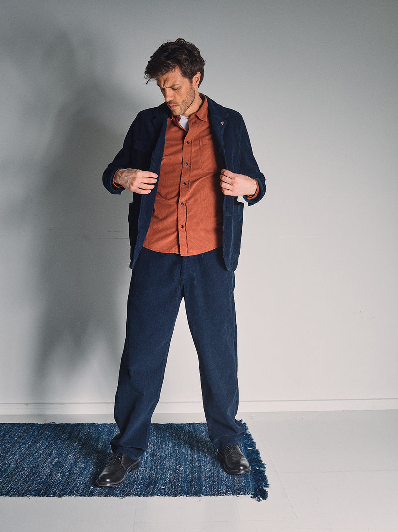 A man stands indoors on a blue rug, wearing a rust shirt beneath the Blue de Gênes Masami Cord Blazer in Navy with matching trousers and black shoes, looking down as he adjusts his corduroy blazer.