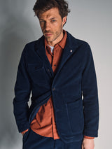 A man with tousled brown hair wears the Blue de Gênes Masami Cord Blazer in navy over a rust button-up, standing against a plain light gray background and looking at the camera with a slight squint.