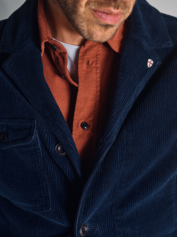 A man wears a navy Masami Cord Blazer by Blue de Gênes over a rusty orange shirt, with part of his face visible. A small pin featuring a red cross is attached to the lapel of the corduroy blazer.