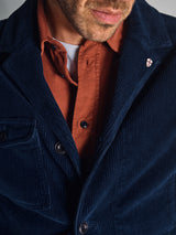 A close-up of a man wearing the Blue de Gênes Zamboni Velu Shirt in Henna under a dark blue corduroy blazer, highlighting the lapel, buttons, and a small pin with a red cross on a white background.