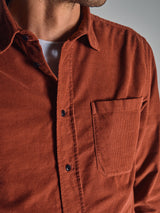 A close-up shows a person wearing the Zamboni Velu Shirt - Henna by Blue de Gênes, a rust-colored baby corduroy shirt with a chest pocket, partially unbuttoned over a white undershirt. Only the lower face and torso are visible.