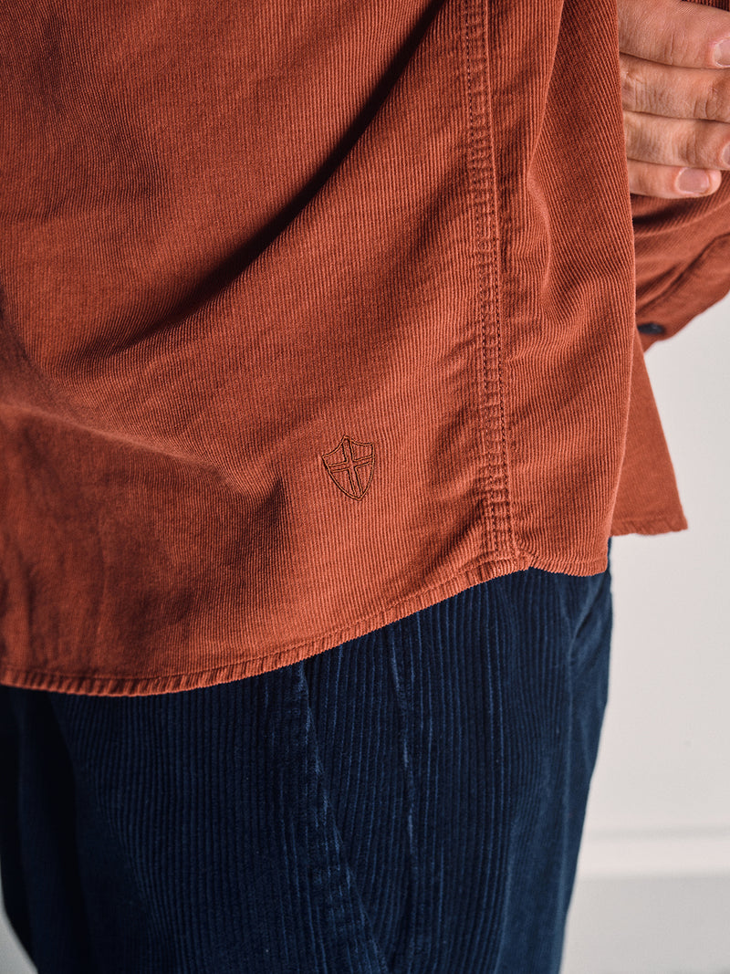 A person wears the Blue de Gênes Zamboni Velu Shirt in Henna, a rust-toned baby corduroy shirt with a small shield logo, paired with dark blue Blue de Gênes corduroy pants and stands with one hand partially visible at their side.