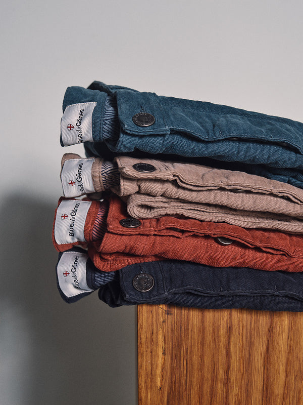 Four pairs of Blue de Gênes BGpaulo Ottawa Trousers in blue, beige, red, and navy are stacked on wood. Made in Italy from dobby corduroy, each features visible buttons and branded waistband labels.